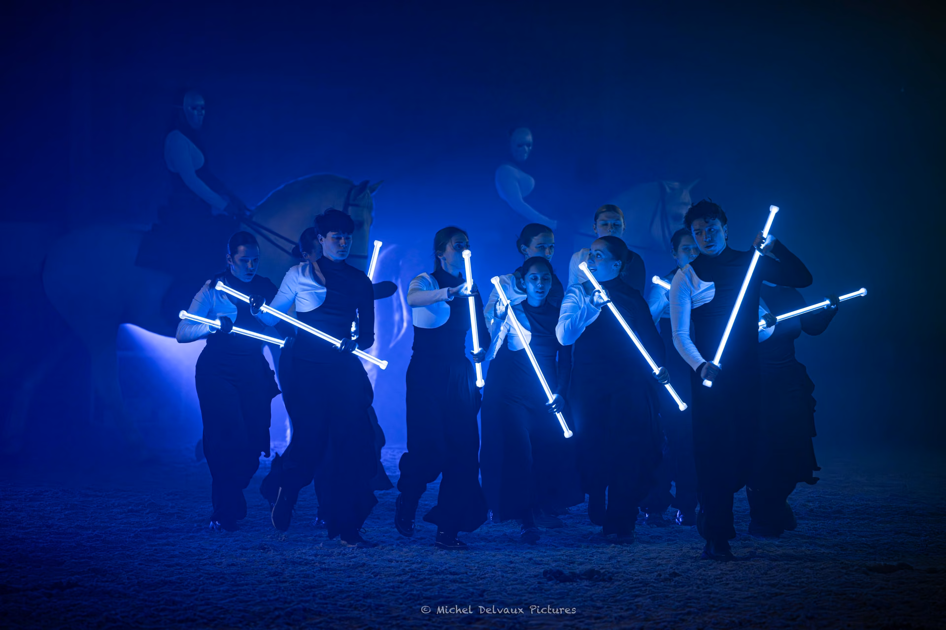 Un groupe d’artistes tient des bâtons lumineux sur une scène éclairée en bleu, devant des projections de chevaux.