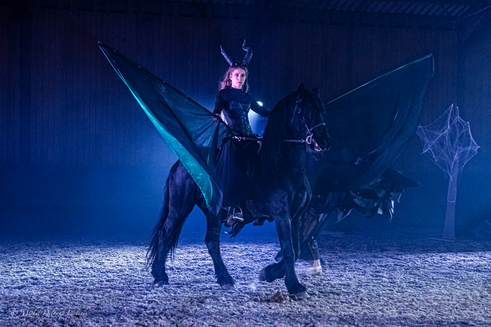 Maléfique : artiste coiffé de cornes sur un cheval noir, déployant une cape verte dans une lumière bleue.
